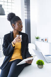 Young female Afro professional looking away holding glass of water and digital tablet while sitting on desk - GIOF11139