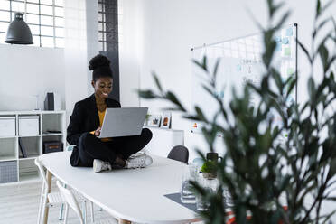 Smiling Afro businesswoman using laptop while sitting cross-legged on desk in office - GIOF11143