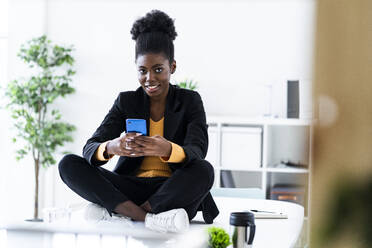 Lächelnde junge Afro-Frau, die im Schneidersitz auf dem Schreibtisch sitzend mit ihrem Handy Textnachrichten verschickt - GIOF11148