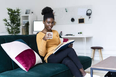 Afro-Frau liest ein Buch und sitzt mit einer Kaffeetasse auf dem Sofa im Wohnzimmer - GIOF11150