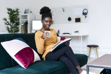 Afro woman reading book while sitting with coffee cup on sofa in living room - GIOF11150