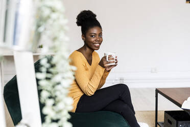 Smiling woman holding coffee cup while sitting on sofa at home - GIOF11151