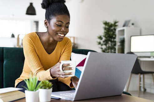 Glückliche Afro-Frau, die eine Tasse hält, während sie einen Laptop zu Hause benutzt - GIOF11154