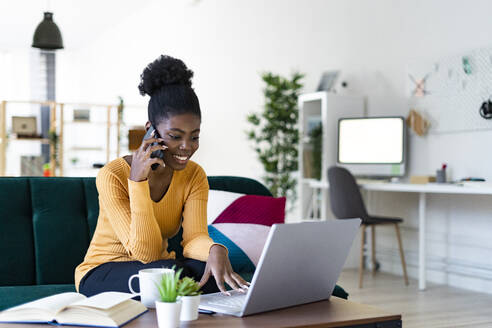 Smiling young woman talking on mobile phone while using laptop in living room - GIOF11156