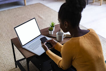 Afro-Frau, die ein Smartphone benutzt, während sie zu Hause am Laptop sitzt - GIOF11157