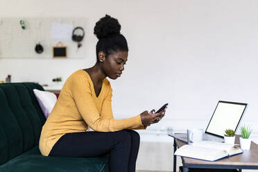 Junge Afro-Frau, die ein Mobiltelefon benutzt, während sie zu Hause im Wohnzimmer sitzt - GIOF11158