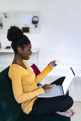 Lächelnde Afro-Frau mit Laptop für Online-Shopping im Wohnzimmer zu Hause - GIOF11162