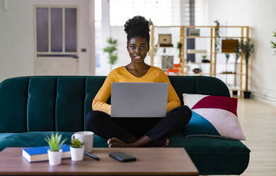 Junge Afro-Frau, die einen Laptop benutzt, während sie im Schneidersitz auf dem Sofa im Wohnzimmer zu Hause sitzt - GIOF11164