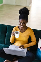 Lächelnde Afro-Frau, die einen Laptop benutzt und eine Kaffeetasse im Wohnzimmer zu Hause hält - GIOF11166