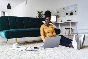 Young Afro woman using laptop while sitting on carpet against sofa in living room at home - GIOF11167