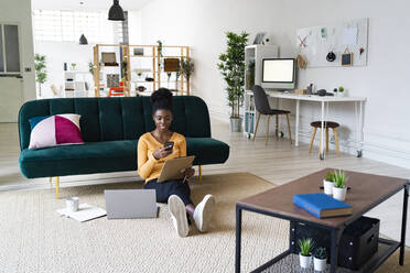 Young woman using smart phone while holding clipboard sitting against sofa in living room at home - GIOF11168