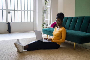 Lächelnde junge Afro-Frau sitzt mit Laptop auf dem Teppich vor dem Sofa im Wohnzimmer zu Hause - GIOF11170