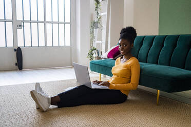 Smiling young Afro woman sitting with laptop on carpet against sofa in living room at home - GIOF11170