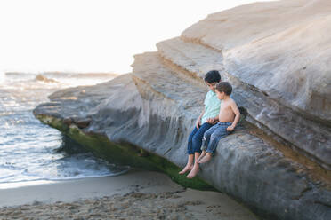 Brüder, die auf einem großen Felsen am Strand sitzen und sich an den Händen halten. - CAVF92524