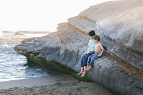 Brüder, die auf einem großen Felsen am Strand sitzen und sich an den Händen halten. - CAVF92524