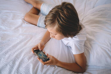 Boy in White Checks plays on his phone from a Hotel Room Bed. - CAVF92538