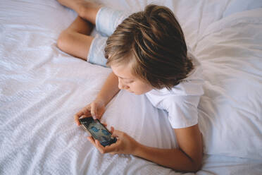 Boy in White Checks spielt auf seinem Handy aus einem Hotelzimmerbett. - CAVF92538