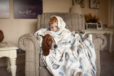 Boy 3-4 years old cuddles with dog in large blanket sitting in chair - CAVF92544
