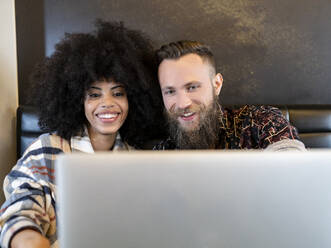 Zufriedenes multiethnisches Paar von Freiberuflern, die an einem Tisch mit Laptop sitzen und gemeinsam an einem Projekt in einem Café arbeiten - ADSF20660