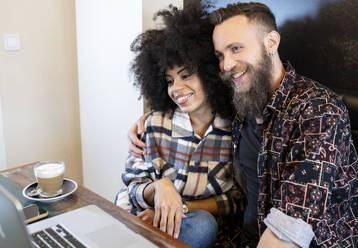 Zufriedenes multiethnisches Paar von Freiberuflern, die an einem Tisch mit Laptop sitzen und gemeinsam an einem Projekt in einem Café arbeiten - ADSF20661