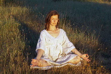 Serene female in dress sitting in Lotus pose with mudra hands in field and meditating while doing yoga with closed eyes in summer - ADSF20665