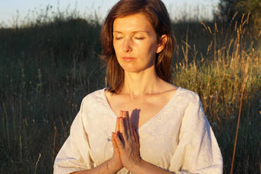 Serene female in dress sitting with prayer hands in field and meditating while doing yoga with closed eyes in summer - ADSF20666