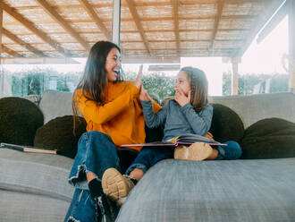 Cheerful female teenager and sibling having fun while spending time on cozy sofa and looking at each other at home - ADSF20668