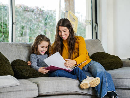 Zufriedener weiblicher Teenager, der die Lehrbuchseite betrachtet, während er Zeit mit seinem Geschwisterchen auf der Couch zu Hause verbringt - ADSF20670