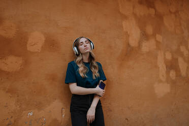 Young mindful blonde female with closed eyes listening to song in wireless headset on brown background in city - ADSF20692