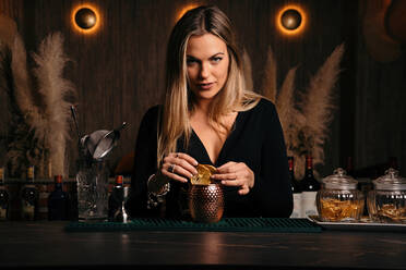 Selbstbewusste attraktive junge Barkeeperin mit langen blonden Haaren in stilvollem Outfit dekoriert Cocktail mit Zitronenscheiben beim Stehen an gezählt in stilvoller Bar - ADSF20700
