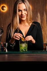 Selbstbewusste junge Barkeeperin mit langen blonden Haaren in stilvollem Outfit dekoriert Cocktail mit Zitronenscheiben beim Stehen an gezählt in stilvoller Bar - ADSF20704