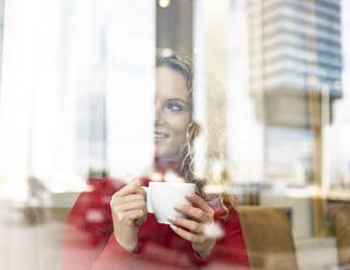 Durch das Fenster einer verträumten Frau, die in einem Café am Tisch sitzt und ein heißes Getränk trinkt, während sie das Wochenende genießt und aus dem Fenster schaut - ADSF20715