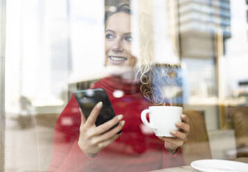 Durch das Fenster einer fröhlichen Frau mit Smartphone und einer Tasse Heißgetränk, die an einem Tisch in einem Café sitzt und wegschaut - ADSF20716