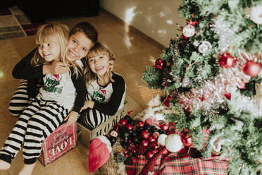 Bruder umarmt Schwestern am Weihnachtsbaum zu Hause - GMLF00948