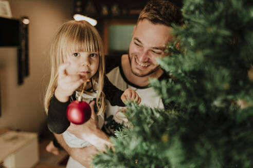 Lächelnder Vater, der ein Mädchen trägt und ein Ornament am Weihnachtsbaum hält - GMLF00955