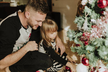 Tochter beim Schmücken des Weihnachtsbaums in der Hocke mit dem Vater zu Hause - GMLF00960