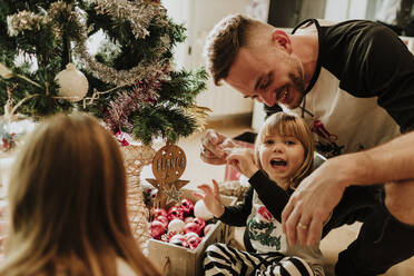 Verspielte Tochter sitzt im Schneidersitz mit der Familie am Weihnachtsbaum zu Hause - GMLF00961