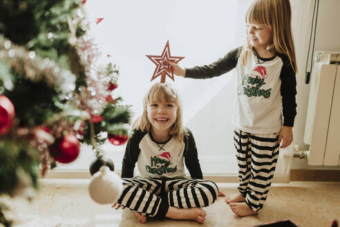 Laughing sisters playing with Christmas decoration against wall at home - GMLF00963