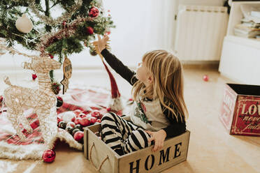Blondes Mädchen berührt den Weihnachtsbaum, während sie im Schneidersitz in einer Schachtel zu Hause sitzt - GMLF00965