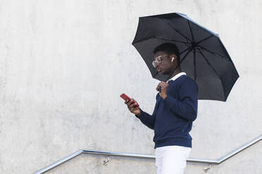 Junger afrikanischer Mann mit Regenschirm hört Musik über In-Ear-Kopfhörer, während er sein Smartphone benutzt - PNAF00611