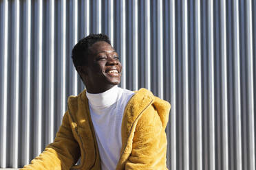Lächelnder afrikanischer Mann in gelber Jacke vor weißer Wand, der wegschaut - PNAF00620