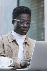 Junger Mann mit Brille benutzt Laptop in einem Straßencafé - PNAF00621