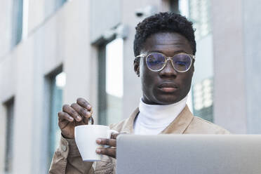 Junger Mann, der in einem Straßencafé Kaffee umrührt und auf seinen Laptop schaut - PNAF00622