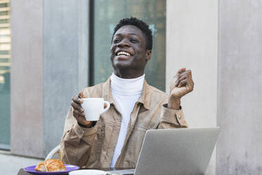 Happy African man with laptop holding coffee cup at sidewalk cafe - PNAF00623