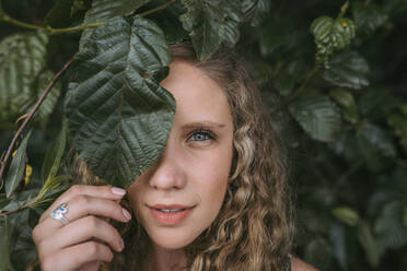 Beautiful blond woman covering eye with leaf in park - DSIF00323