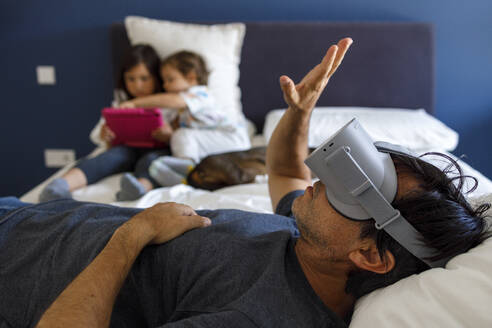Mann mit Virtual-Reality-Simulator gestikuliert, während Geschwister auf dem Bett zu Hause ein digitales Tablet benutzen - IFRF00399