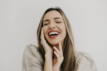 Laughing woman touching cheek with eyes closed against white background - DSIF00335