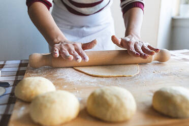 Woman flattening dough with rolling pin on cutting board to make croissants in kitchen - WPEF04014