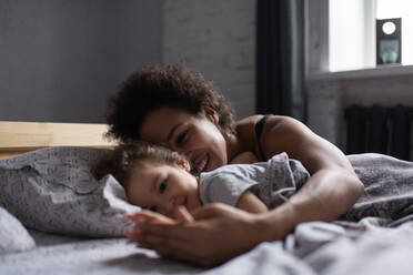 Fröhliche gemischtrassige Mutter und Tochter liegen morgens auf dem Bett - CAVF92683