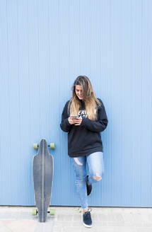 Junge Frau mit Skateboard und Smartphone, während sie auf einem Bein vor einer blauen Wand steht - DAMF00700